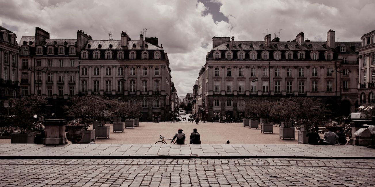 two person sitting on pathway beside beige and white concrete building under white skies