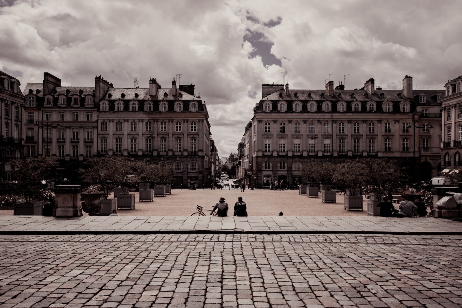 two person sitting on pathway beside beige and white concrete building under white skies